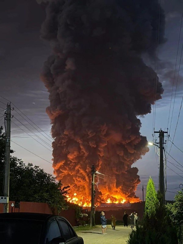 A massive fire erupts behind a fence at dusk, sending a towering plume of thick black smoke into the sky as onlookers gather nearby.