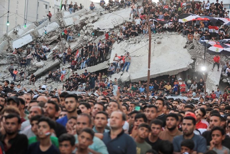 A large crowd gathers at night around a collapsed building, with people sitting atop the rubble and Egyptian flags visible among the onlookers.