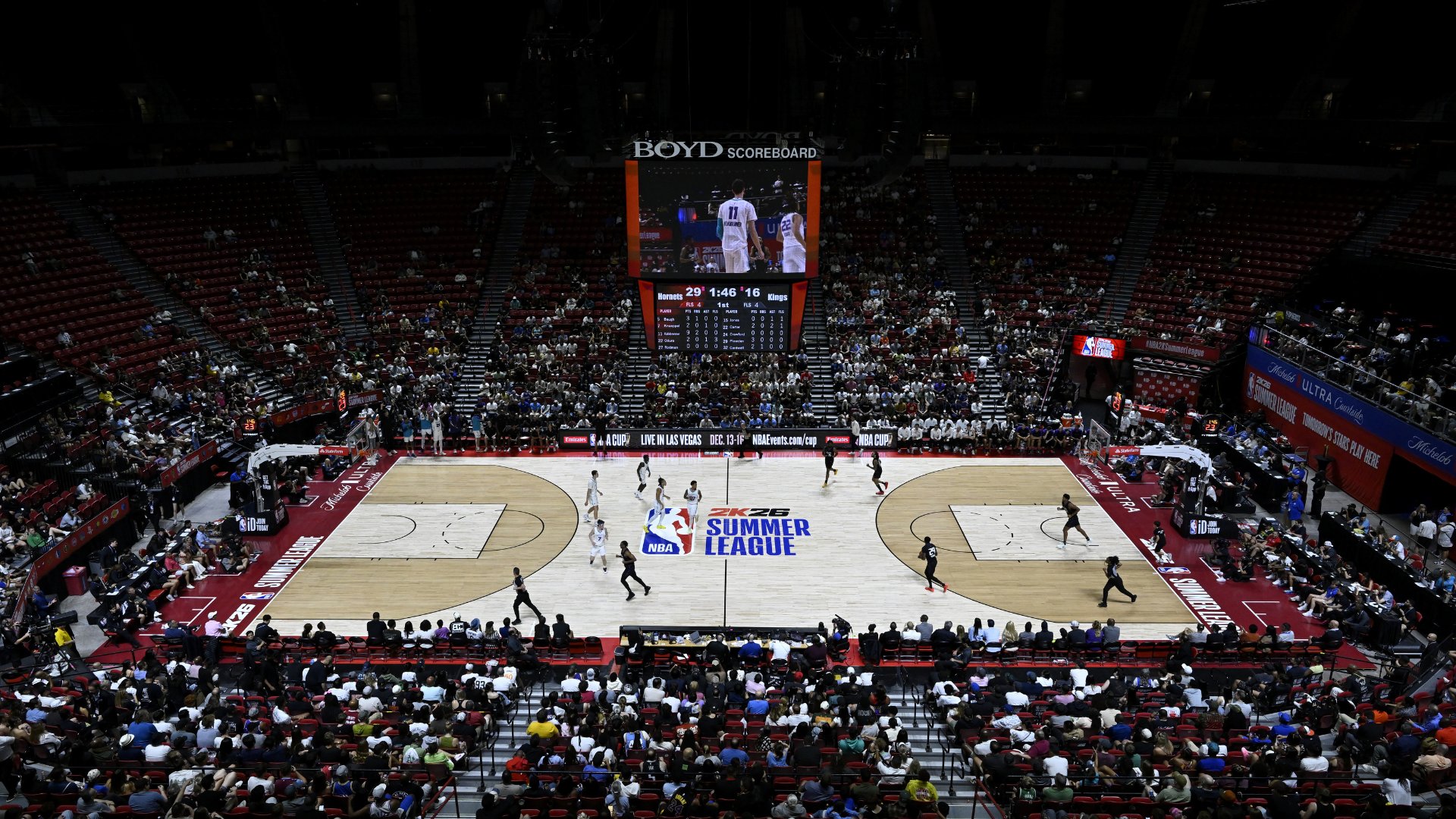 Basketball players compete on a hardwood court at an NBA Summer League game, with a scoreboard overhead showing the Hornets vs. Kings and spectators filling the arena seats.
