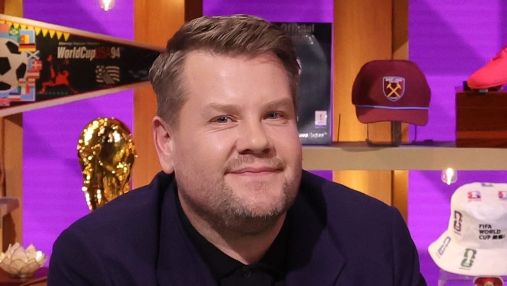 A man in a dark blazer smiles on a purple-lit set decorated with a World Cup trophy, soccer ball, and caps.