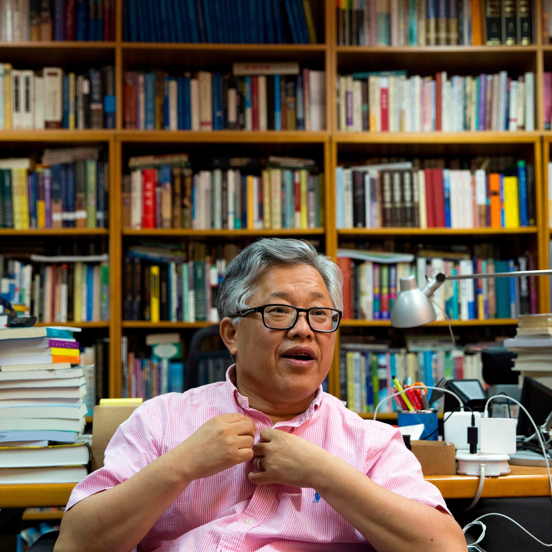 A gray-haired man wearing glasses and a pink shirt sits in front of a large bookshelf, gesturing with his hands while speaking.