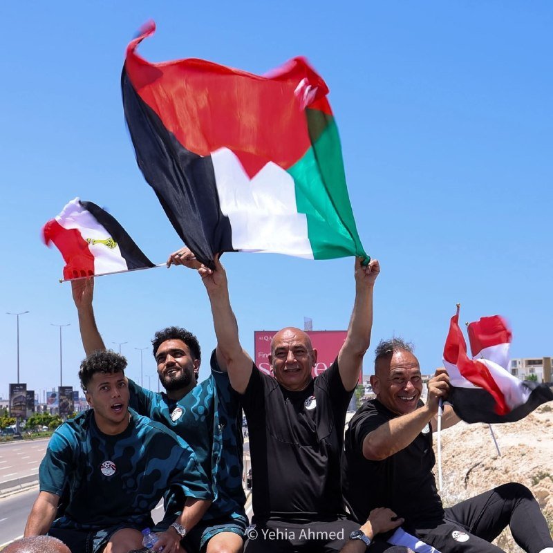 Four men smile while waving red, black, white, and green flags against a clear blue sky outdoors.