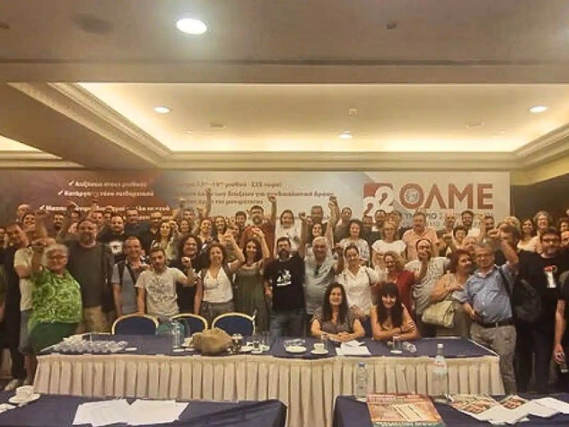 A large group of people gathers behind a long table in a conference room, raising their fists toward the camera in front of a banner displaying "OLME" and the number "20."