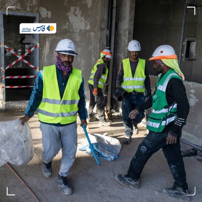 Four construction workers in high-visibility vests and hard hats walk through an unfinished building, carrying bags and tools amid scattered debris.