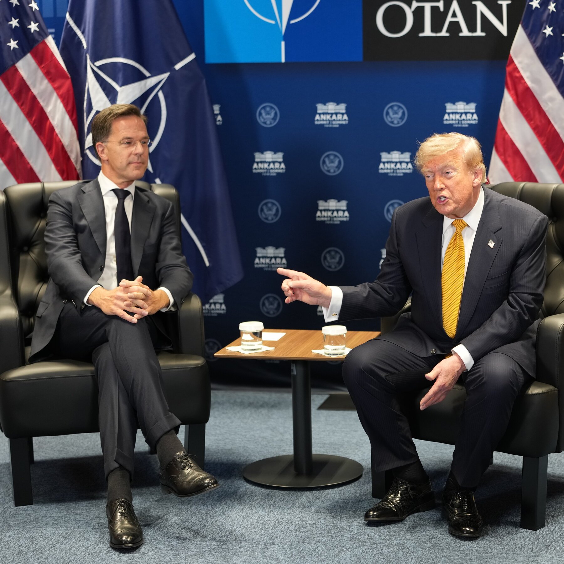 Two men in suits sit in black leather chairs at a NATO-branded summit, with American flags and an "ANKARA" backdrop visible behind them.