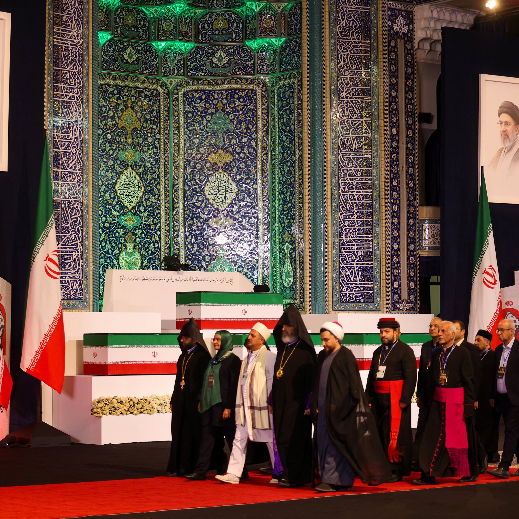 A large crowd waves Iranian flags and holds up signs near a yellow construction barrier, with armed personnel standing on an elevated platform in front of a multi-story unfinished building.