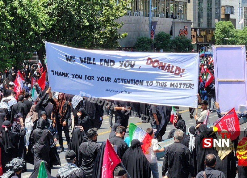 A crowd marches down a city street holding a large banner reading "WE WILL END YOU, DONALD! THANK YOU FOR YOUR ATTENTION TO THIS MATTER!" alongside various flags.