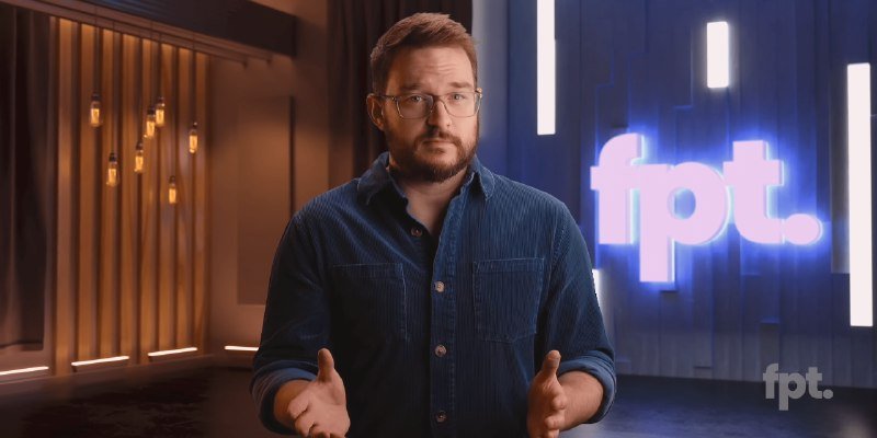 A bearded man wearing glasses and a blue corduroy shirt gestures with open hands in a studio setting, with an illuminated "fpt." sign displayed behind him.