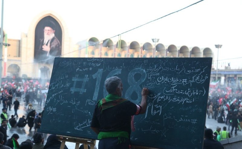 A man writes "#118" and Persian text on a chalkboard before a crowd gathered in a large plaza featuring a prominent portrait of a bearded cleric on a building.