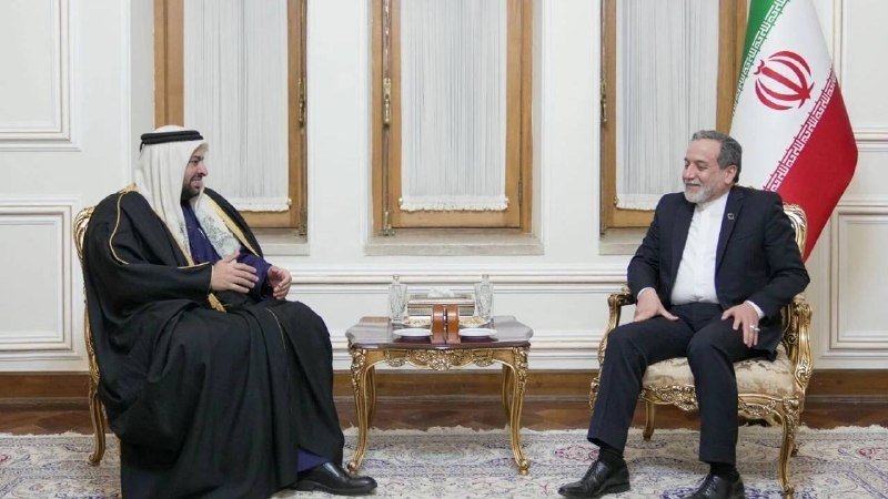 Two men sit facing each other in ornate chairs across a small table, one in traditional Gulf attire beside a green flag, the other in a dark suit beside the Iranian flag.