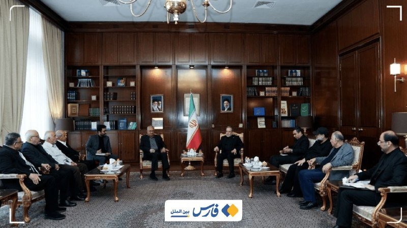 A group of men sit in a wood-paneled meeting room with bookshelves, seated around low tables displaying an Iranian flag and teacups in the center.