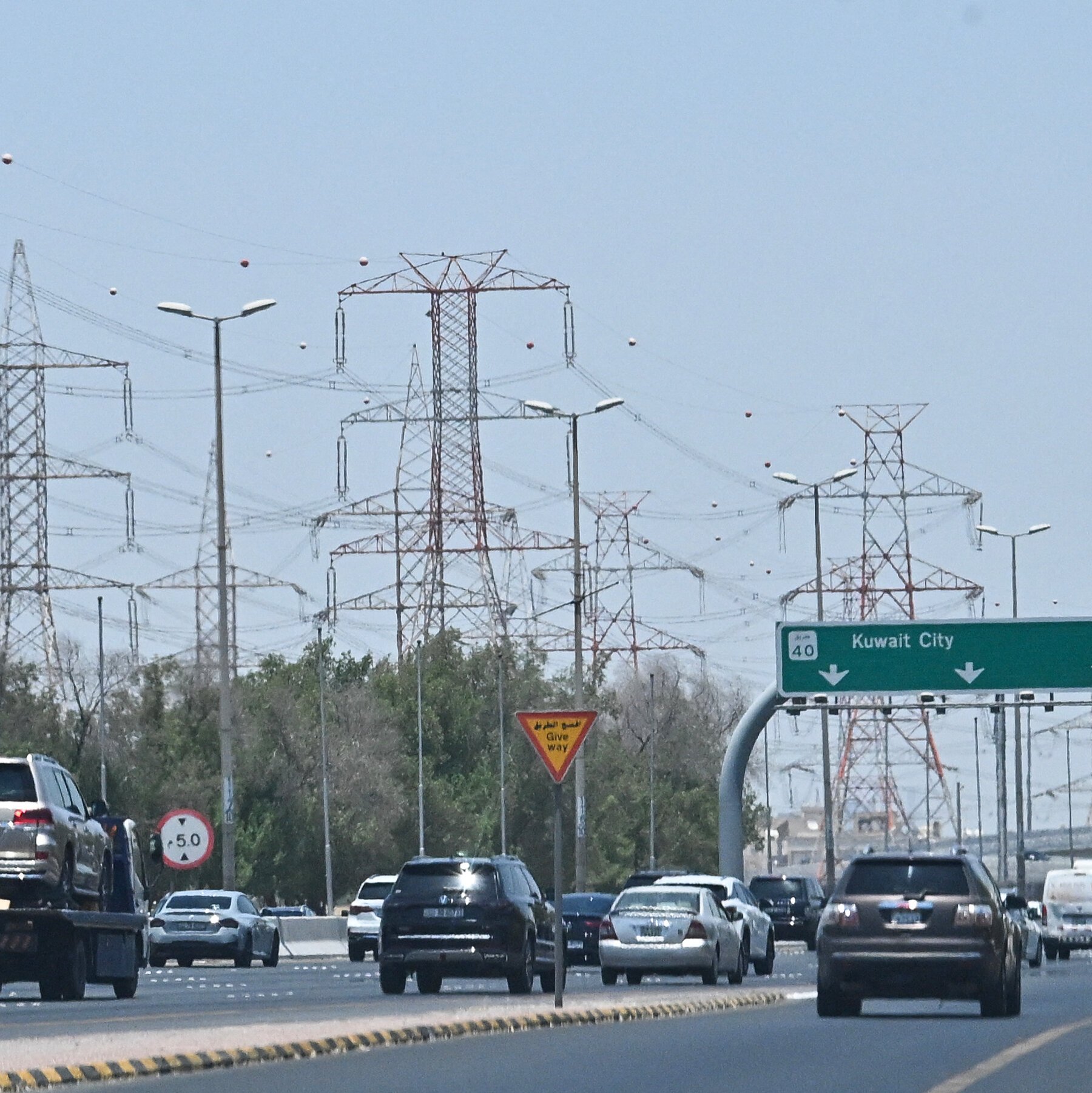 Vehicles travel along a multi-lane highway beneath a network of electrical transmission towers, with a green sign reading "Kuwait City" visible overhead.