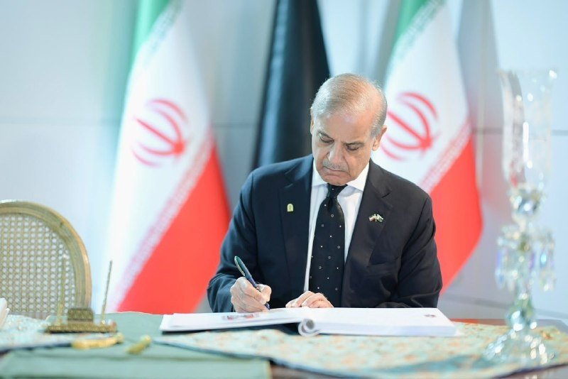 A man in a dark suit signs a document at a desk, with Iranian flags visible in the background.