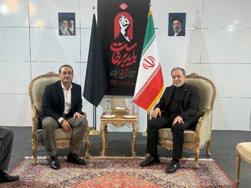 Two men in formal attire — one in traditional Arab dress, the other in a dark suit — sit in ornate chairs across a small table, with an Iranian flag visible behind them.