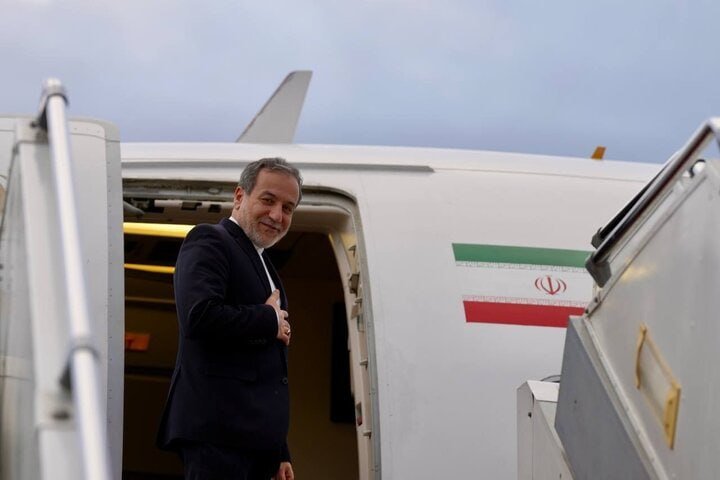 A bearded man in a dark suit stands at the doorway of an aircraft displaying the Iranian flag emblem on its fuselage.