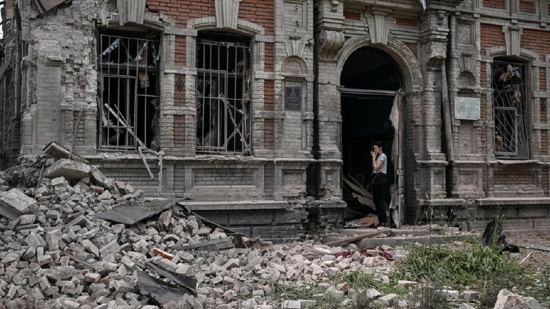 A man talks on a phone in the doorway of a heavily damaged brick building surrounded by a large pile of rubble and shattered glass.