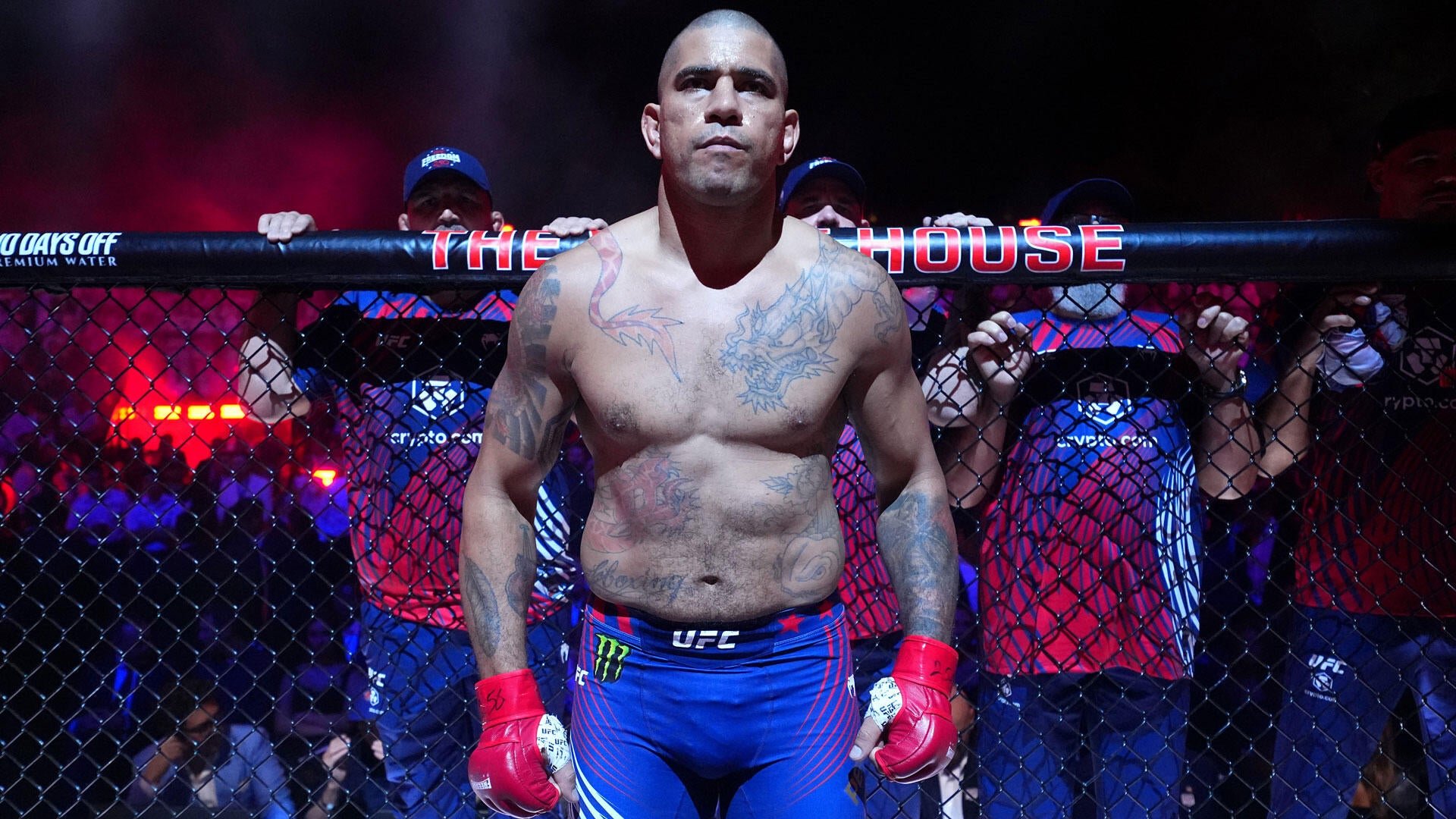 A shirtless, tattooed fighter wearing blue UFC shorts and red gloves stands inside a cage, gripping the fence as officials in blue shirts stand behind him.