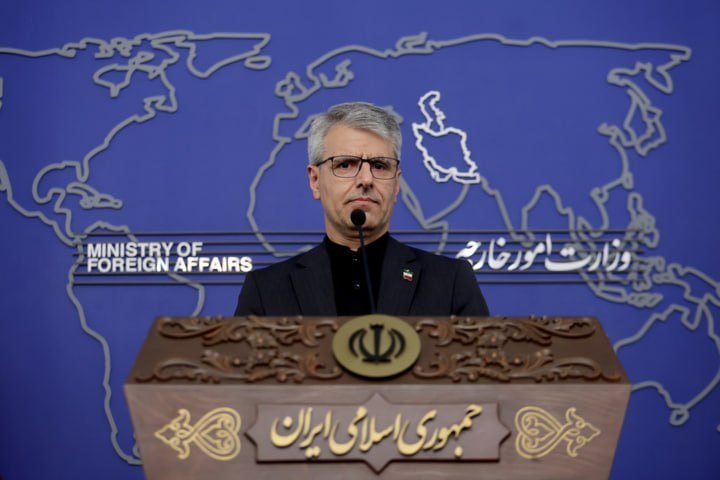 A bespectacled man in a dark suit stands at a podium featuring an emblem and Persian text, with a blue backdrop displaying a world map and "MINISTRY OF FOREIGN AFFAIRS."