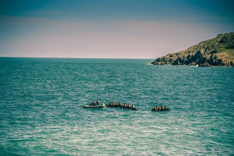 Three small boats carrying groups of people float in turquoise ocean waters near a rocky, vegetated coastline under a clear sky.
