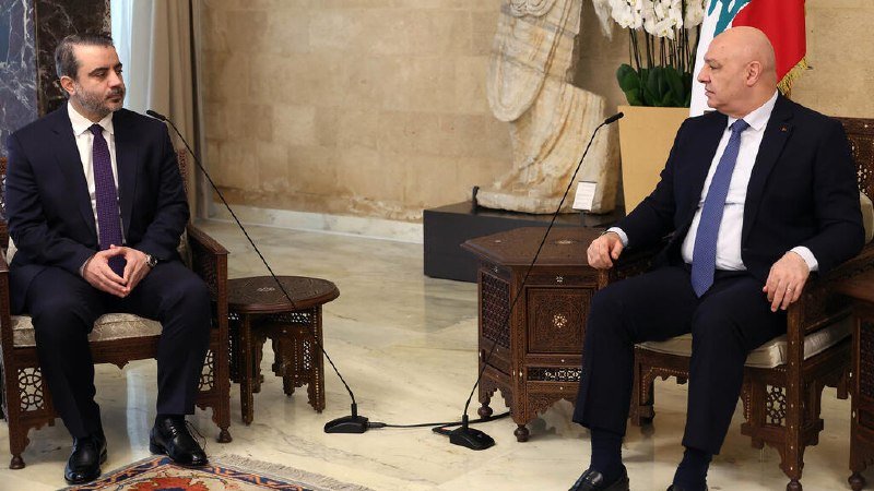 Two men in dark suits sit on a beige sofa in a warmly lit lounge, with the man on the left gesturing while speaking, as Arabic text and a Shaam Network logo appear below.