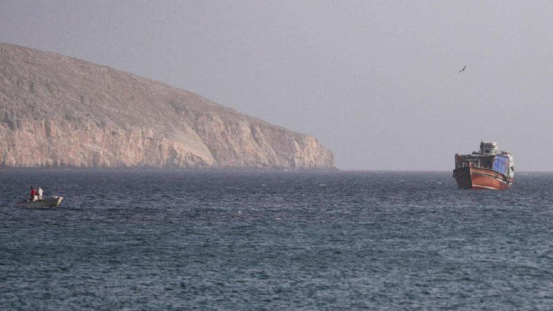 A small boat with two people and a large cargo ship float on the sea near a steep, rocky coastline under a hazy sky.