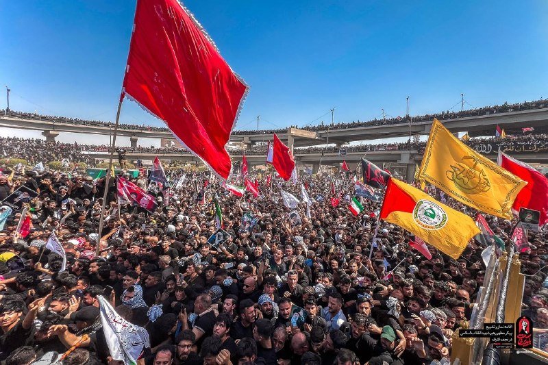 A massive crowd fills a dusty open area beneath an overpass, with mourners waving large red, yellow, and black flags under a clear blue sky.