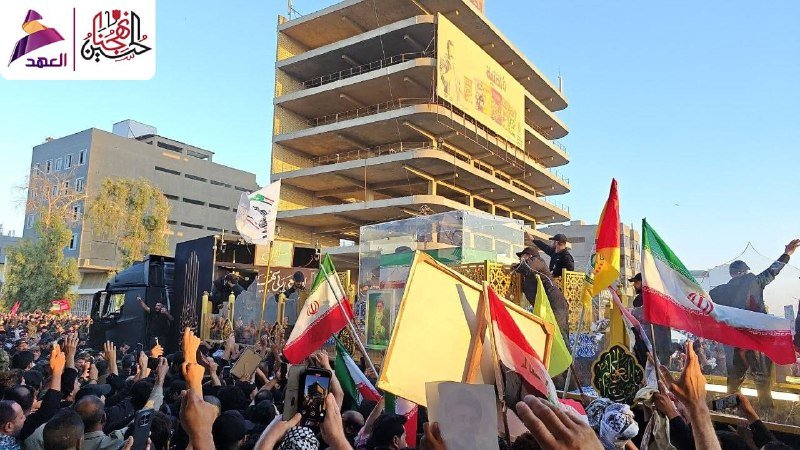 A large crowd waves Iranian flags and posters in front of a yellow-framed casket at an outdoor gathering, with an unfinished multi-story building in the background.