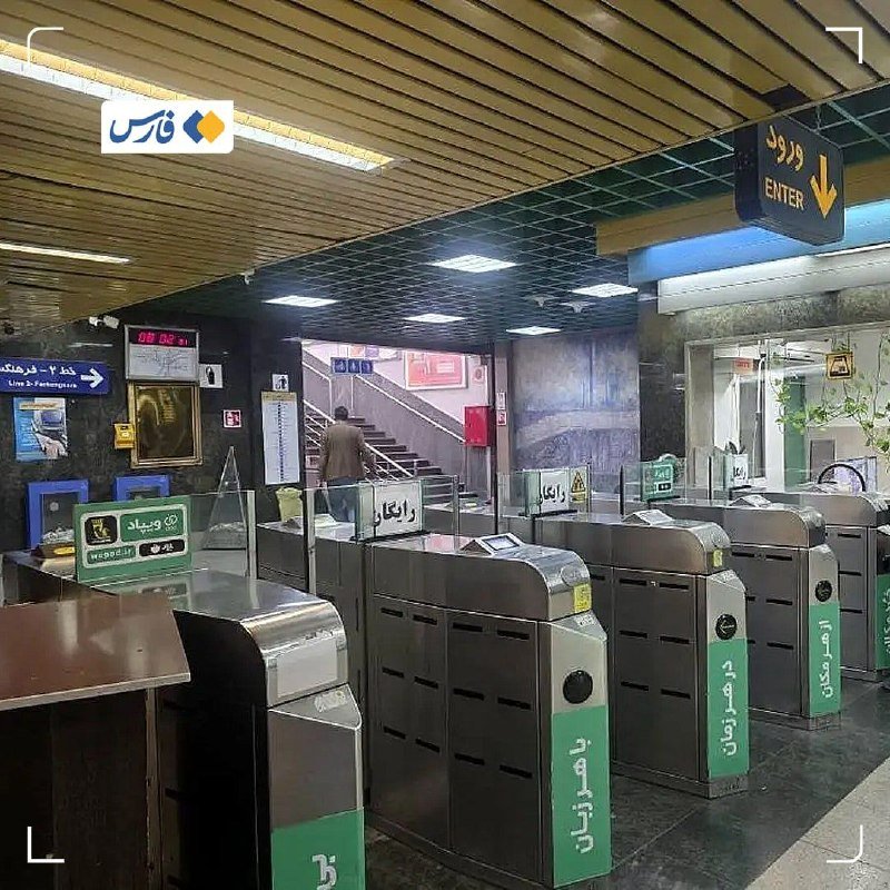 A row of metal turnstiles with green panels stands inside a transit station, with a person ascending a staircase beneath illuminated ceiling lights and bilingual signage.