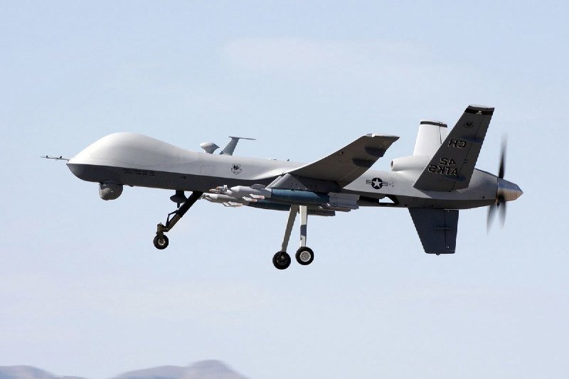 A gray military drone marked "HQ" and "0W CNAF" flies with landing gear deployed against a pale sky above distant mountains.