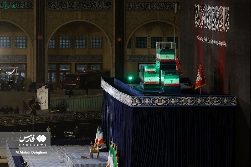 Stacked coffins draped in the Iranian flag sit on an elevated, blue-curtained platform inside a large mosque with arched architecture.