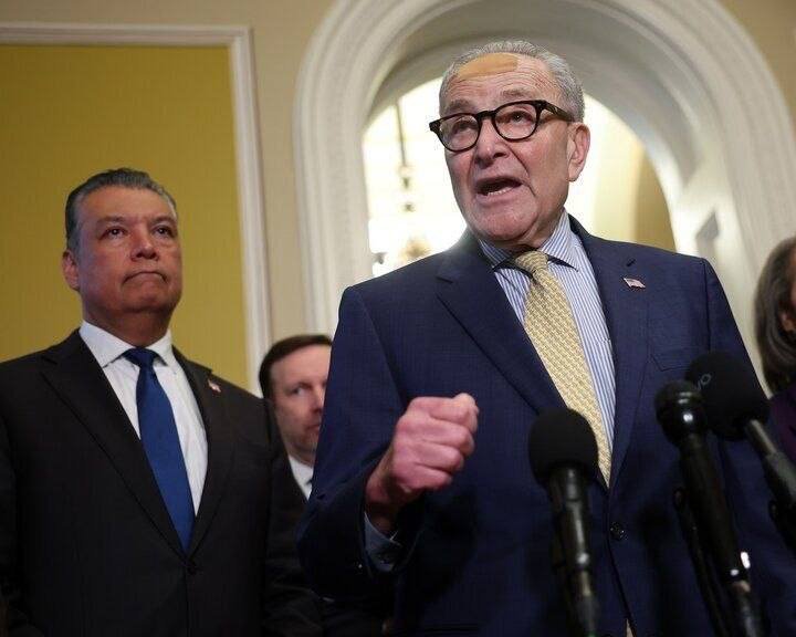 An older man in a blue suit and glasses speaks at microphones with his fist raised, while another man in a dark suit stands behind him in a hallway.