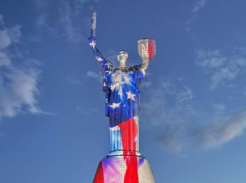 A large motherland-style statue holding a sword and shield is illuminated in red, white, and blue against a partly cloudy sky.