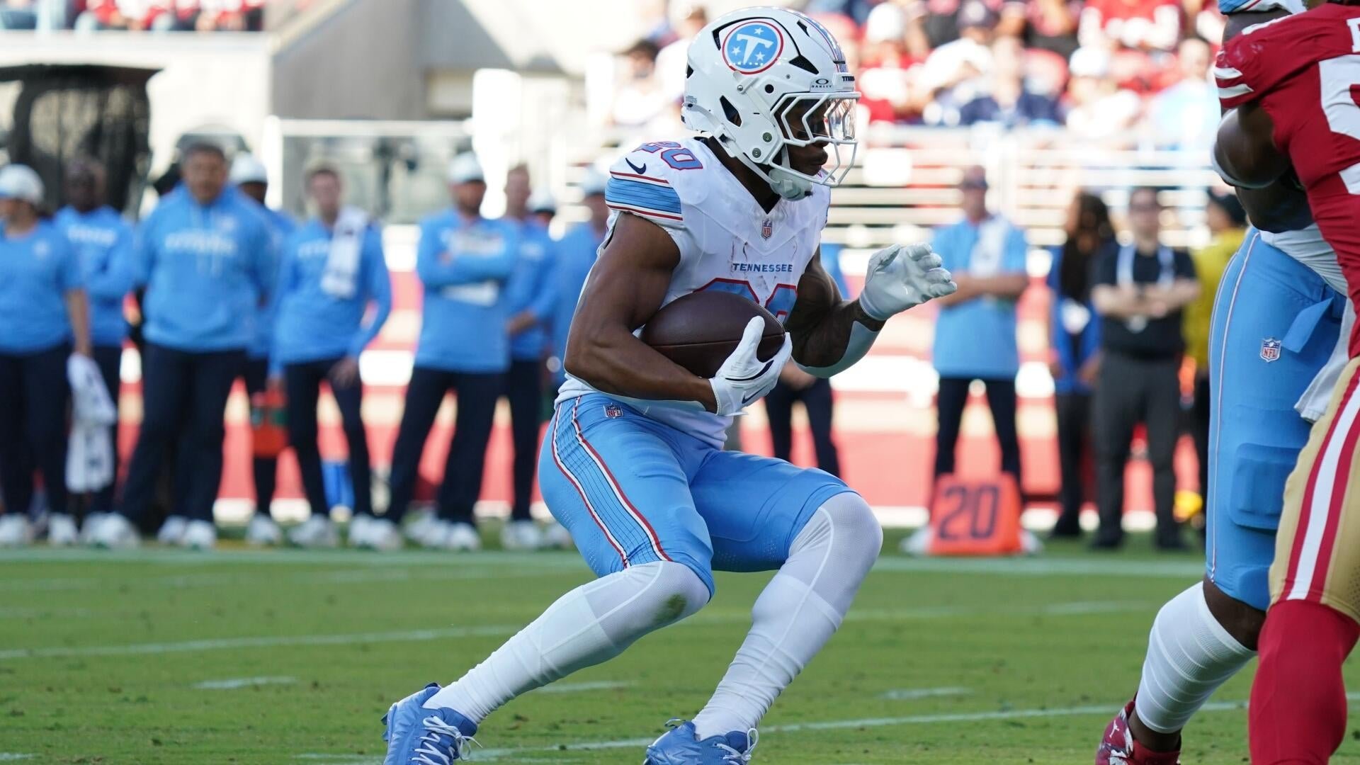 A football player in a white and light blue Tennessee Titans uniform runs with the ball tucked under his arm during a game, with sideline personnel visible in the background.