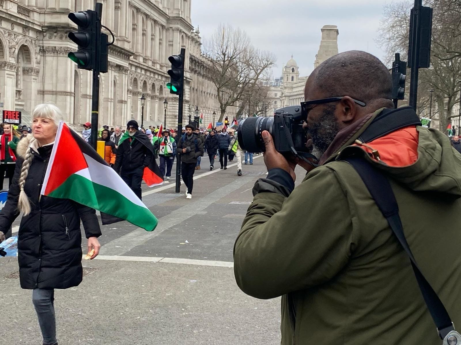 A photographer photographs a woman carrying a Palestinian flag during a march down a city street, with a "FREE PALESTINE" sign visible.