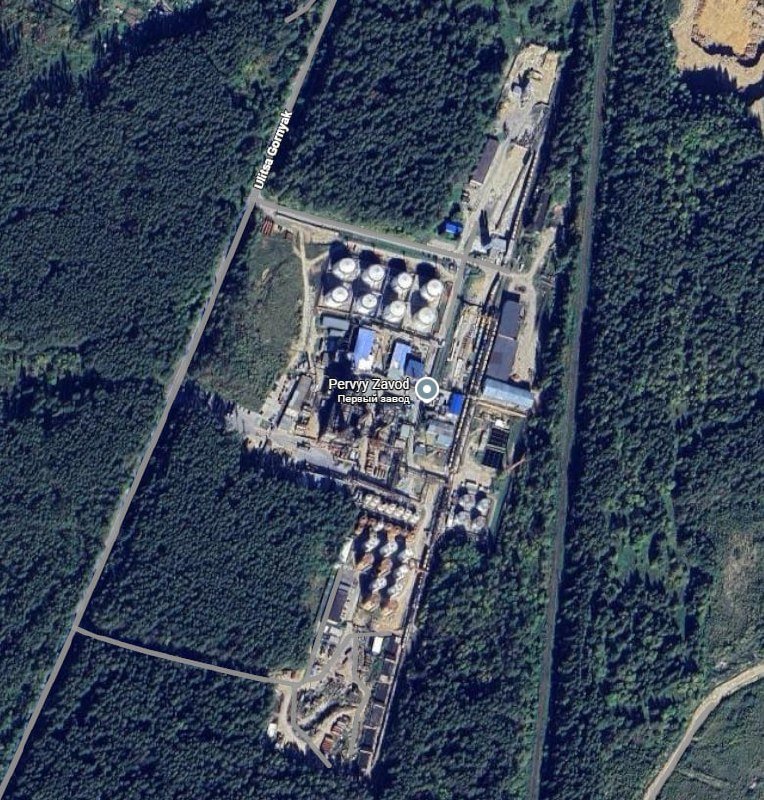 Satellite view shows an industrial facility with storage tanks and buildings labeled "Pervyy Zavod" surrounded by dense forest and intersecting dirt roads.