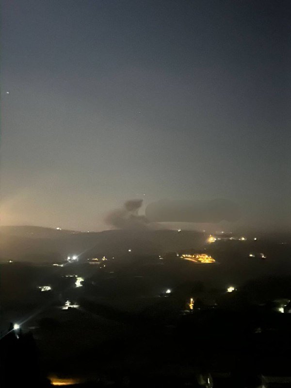 Nighttime view over a valley shows scattered city lights below a thick layer of low clouds or fog covering the distant hills.