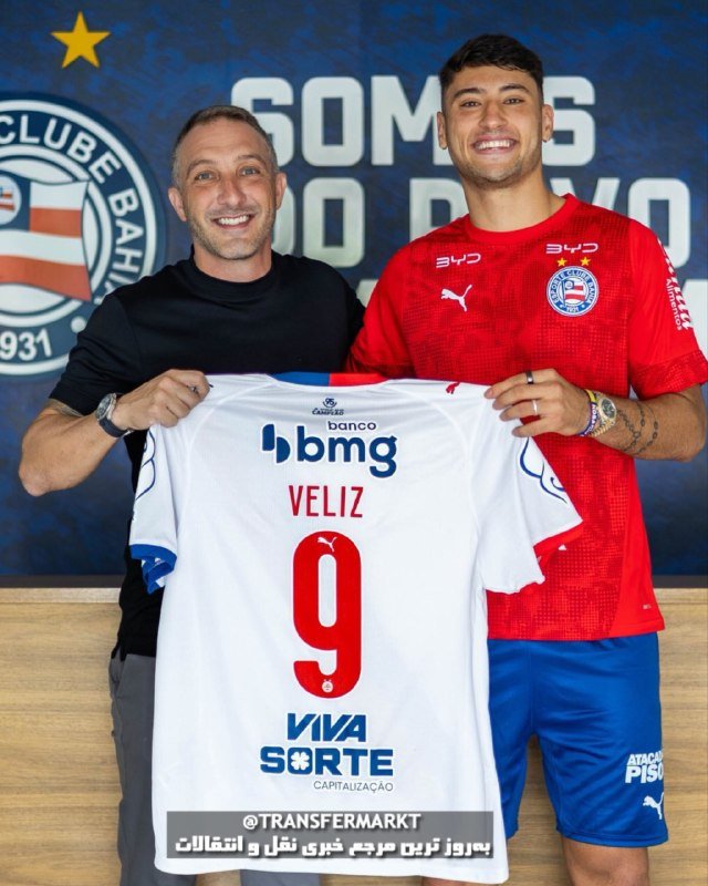 Two men smile while holding up a white jersey reading "VELIZ 9" with sponsor logos, in front of a Bahia club backdrop.
