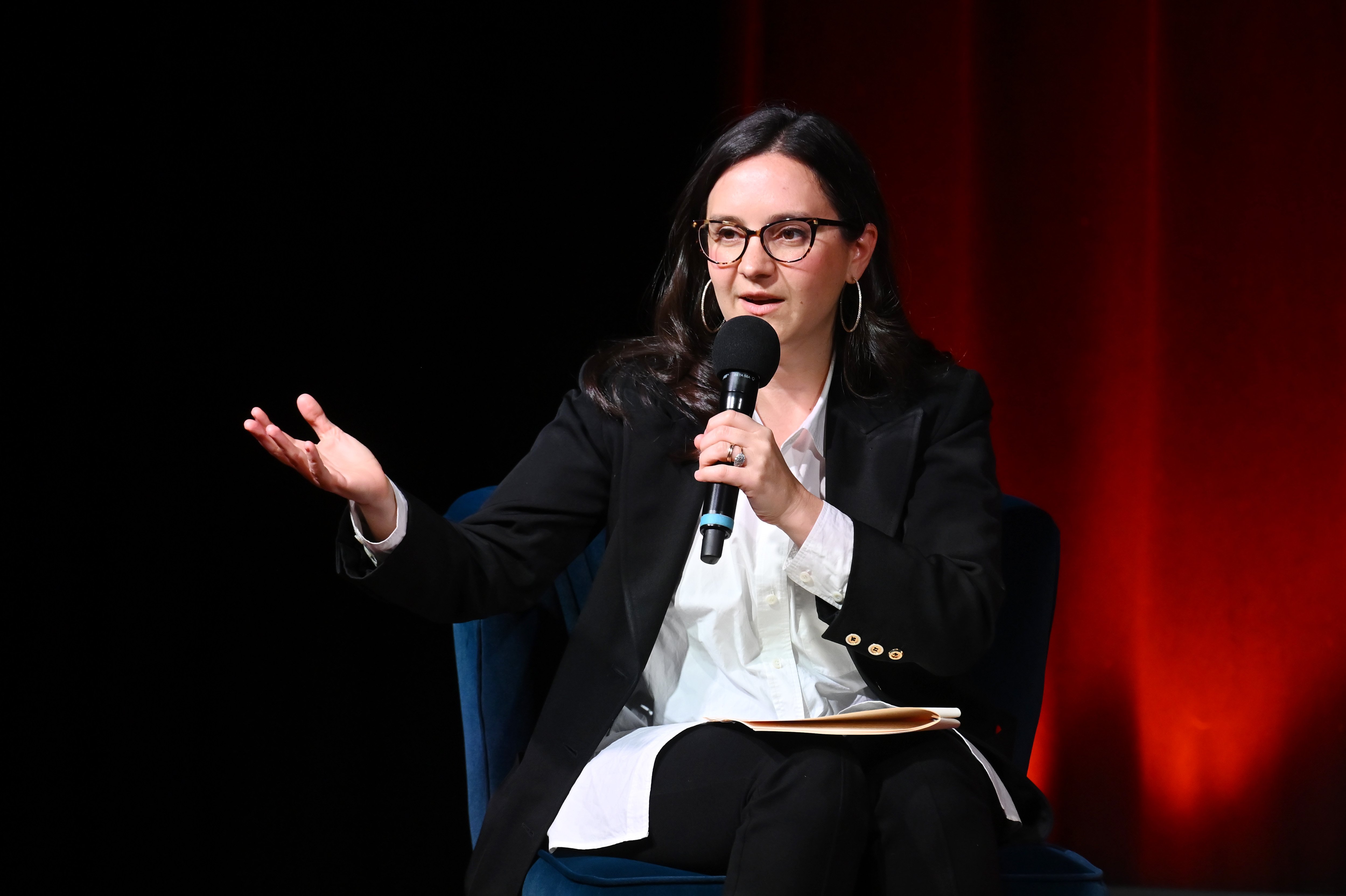A woman with dark hair and glasses, wearing a black blazer over a white shirt, speaks into a microphone while gesturing with one hand, seated against a dark red backdrop.
