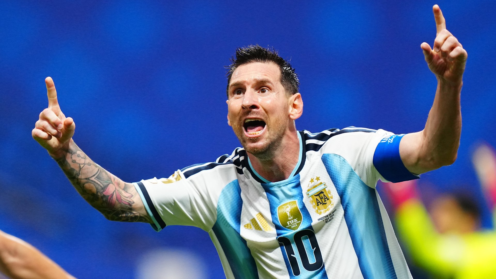 A soccer player wearing an Argentina #10 jersey with tattoos on his right arm raises both index fingers skyward while shouting in celebration against a blue background.