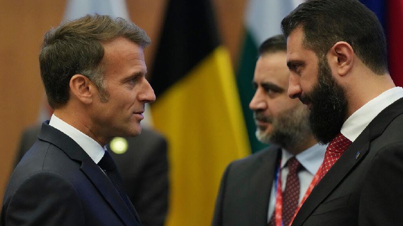 Three men in dark suits converse in profile, with blurred national flags visible in the background.