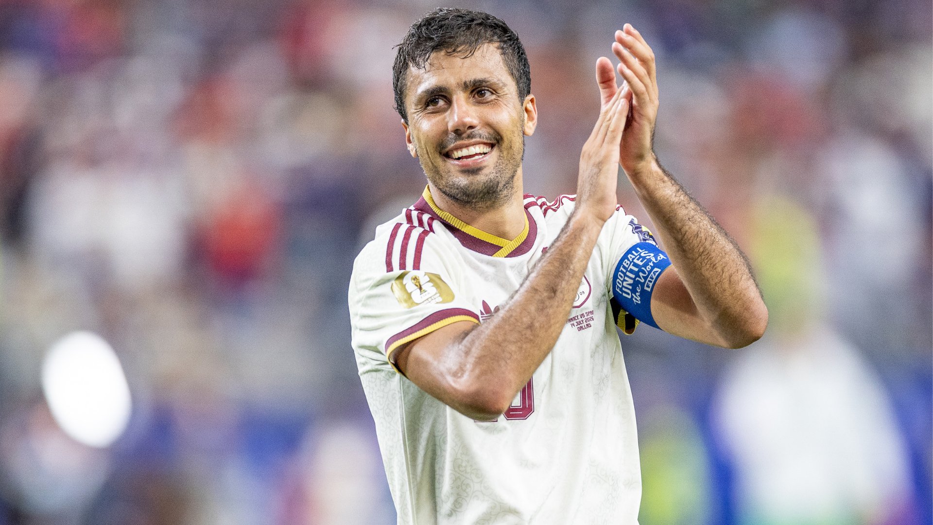A smiling soccer player in a white and maroon jersey claps his hands in front of a blurred crowd, wearing a "Football United the World" armband.