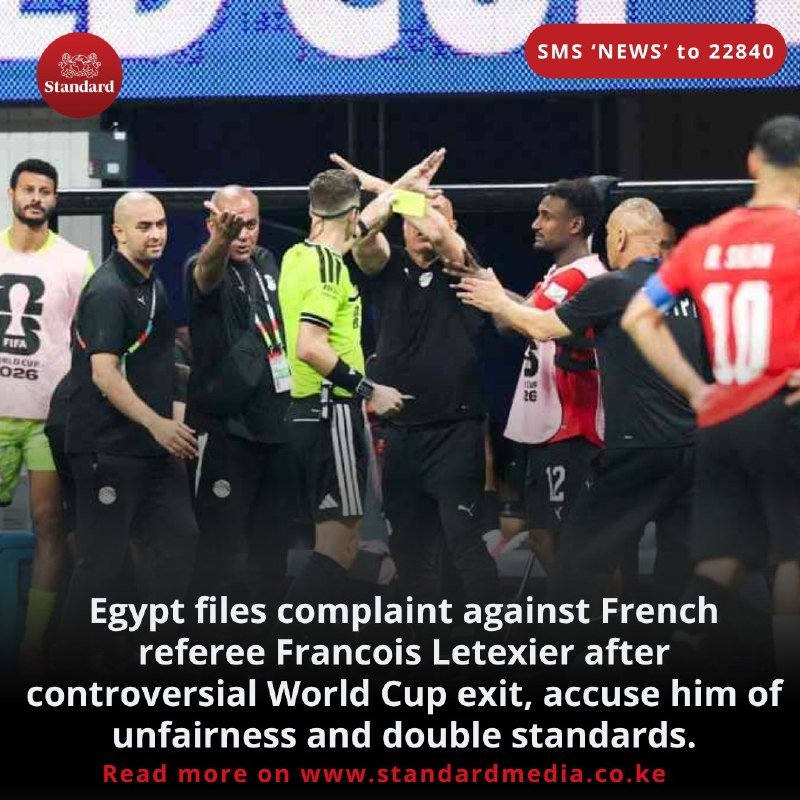 A football referee in a yellow shirt holds up a yellow card while surrounded by coaching staff and players gesturing and arguing on the touchline.