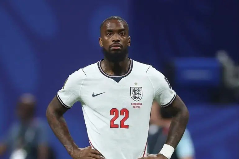 A bearded soccer player wearing a white England away jersey with the number 22 stands with hands on hips against a blurred blue stadium background.