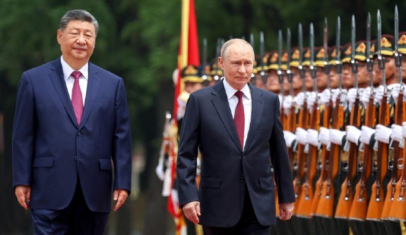 Two men in dark suits walk past a row of uniformed honor guards holding rifles during an outdoor ceremonial reception.