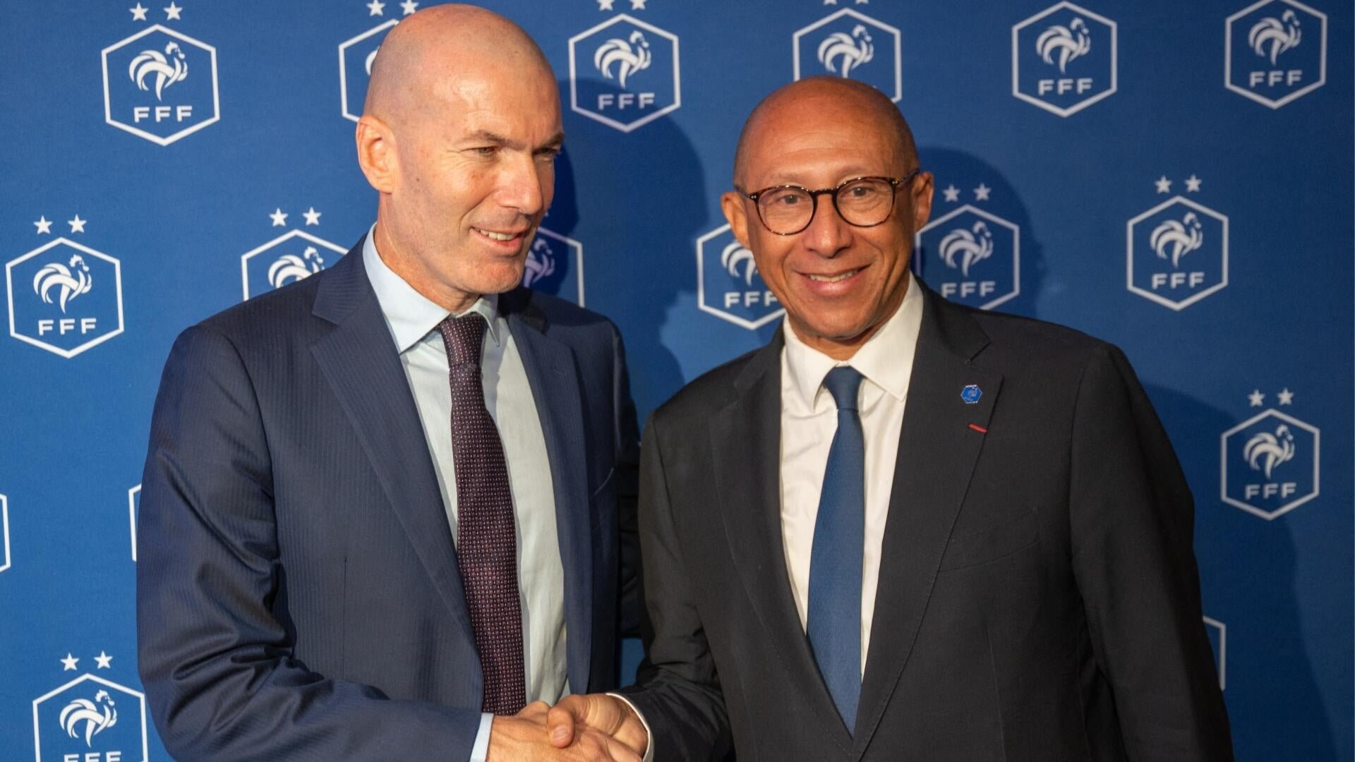Two men in suits smile and shake hands in front of a blue backdrop featuring the FFF (French Football Federation) logo with a rooster emblem.
