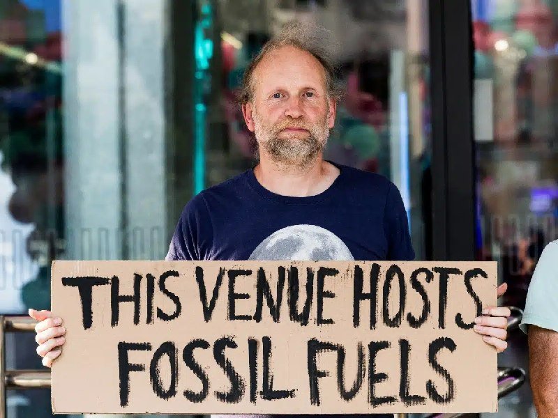 A bearded man in a navy moon-print shirt holds a cardboard sign reading "THIS VENUE HOSTS FOSSIL FUELS" in front of a glass storefront.