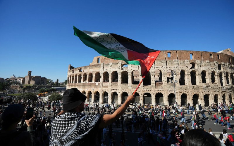 A man wearing a black-and-white keffiyeh scarf and black shirt stands with arms outstretched in front of a heavily damaged concrete building.