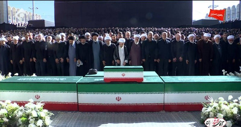 Clerics in turbans and robes stand behind a coffin draped in the Iranian flag, with a large crowd gathered at an outdoor ceremony.
