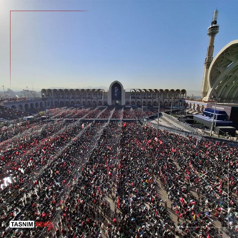 A large crowd fills a plaza in front of an arched building displaying a portrait, with many red flags visible and a tall tower nearby; the Tasnim News logo appears.