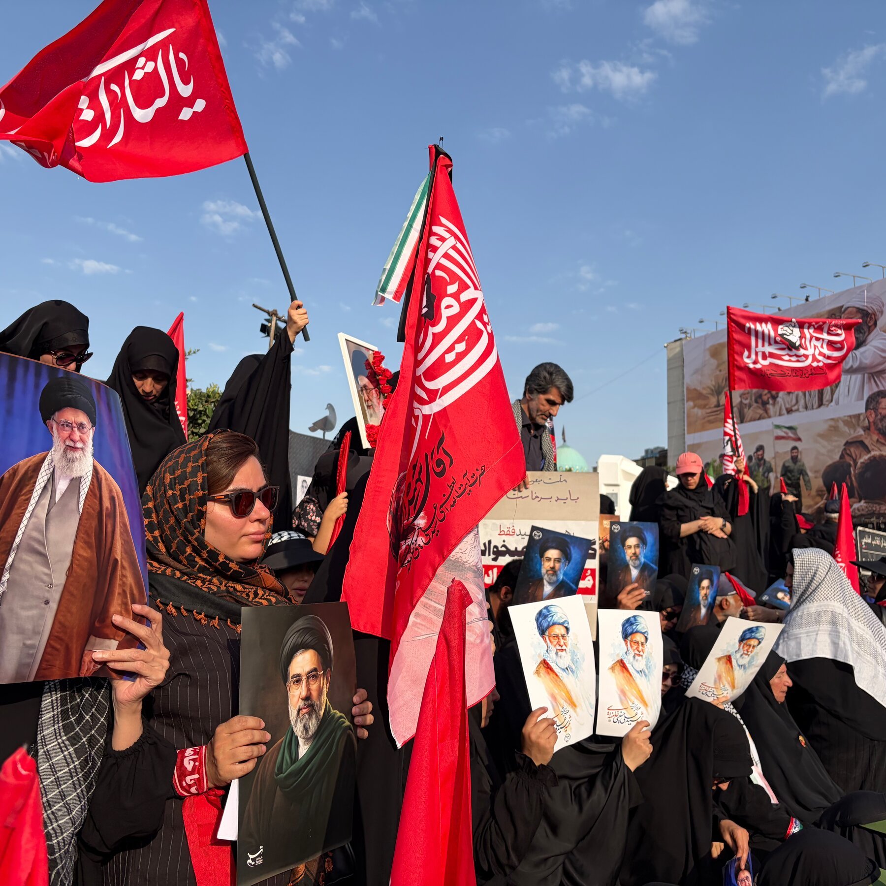 A crowd of people, many in black clothing, hold portraits of bearded men in turbans and large red flags with Arabic script during an outdoor daytime gathering.